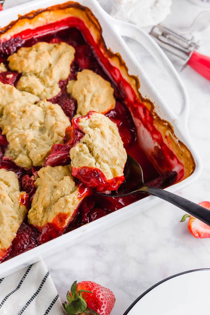 A photo of a baking sheet with strawberry cobbler topped with a spoon scooping out gluten free vegan biscuits.