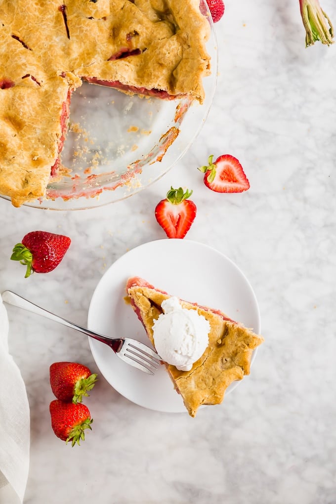 A piece of rhubarb strawberry pie on a plate with a fork and a larger gluten free pie. 