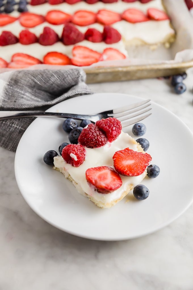 A piece of American Flag Cookie Cake on a plate topped with raspberries, blueberries and strawberries with the full gluten free cookie cake in the background. 