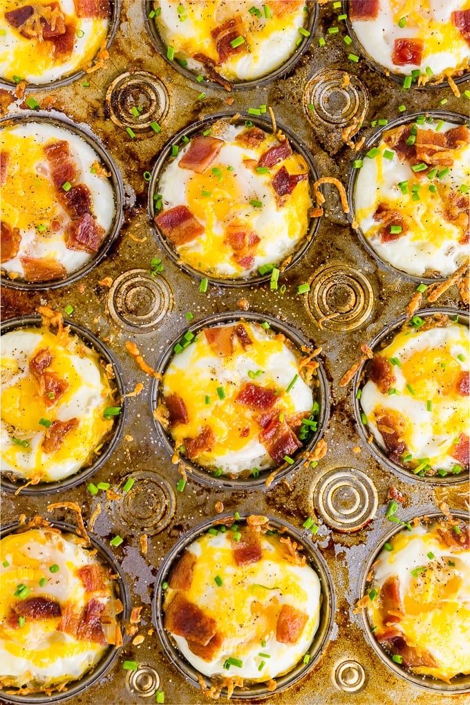 A photo of a muffin tin filled with gluten free hash brown breakfast egg cups topped with bacon, chives and cheddar cheese.