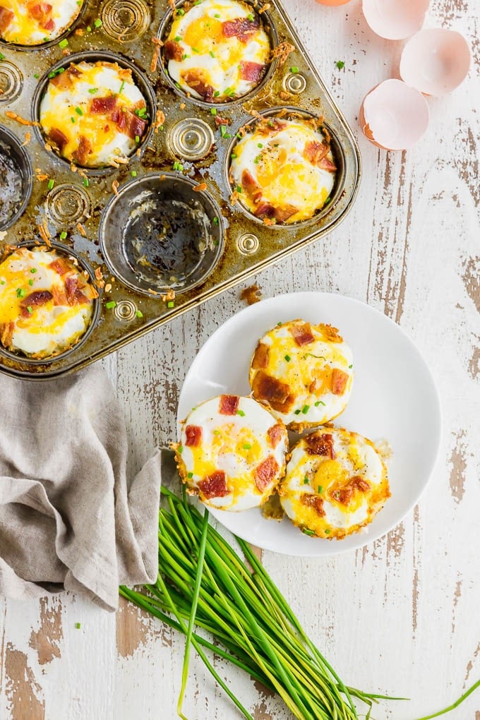 A photo of a muffin tin filled with hash brown egg cups with three egg cups on a plate with a bunch of chives. 