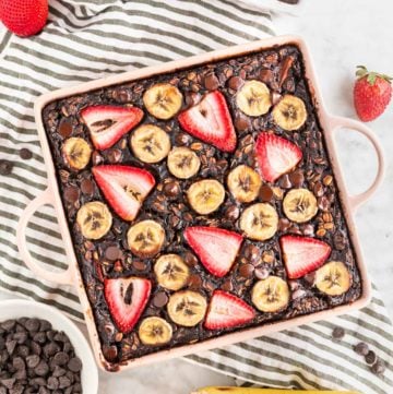 A photo of a baking dish with chocolate strawberry banana baked oatmeal in it with sliced bananas and strawberries on top and a bowl of chocolate chips next to it.