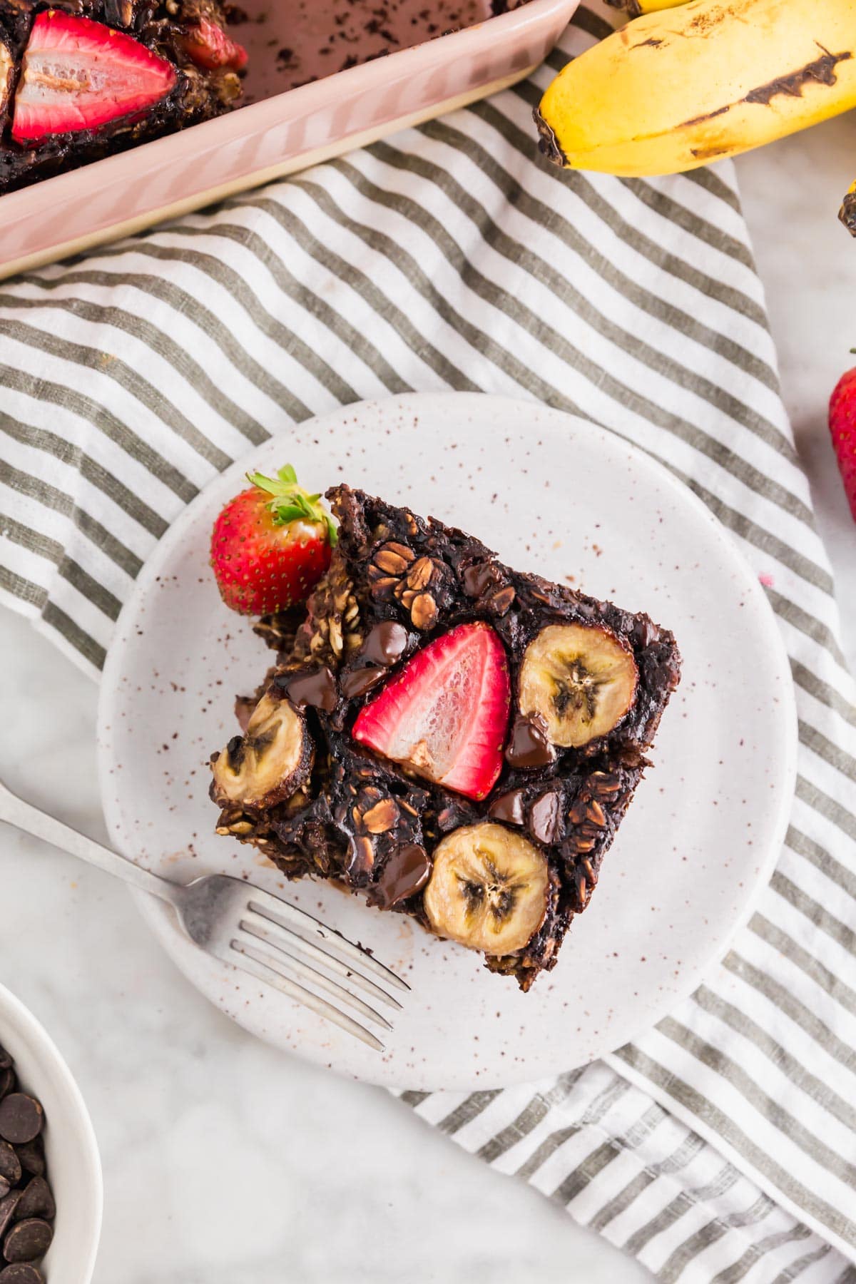 A photo of a plate with a slice of gluten-free chocolate baked oatmeal topped with strawberries and bananas with a fork. 