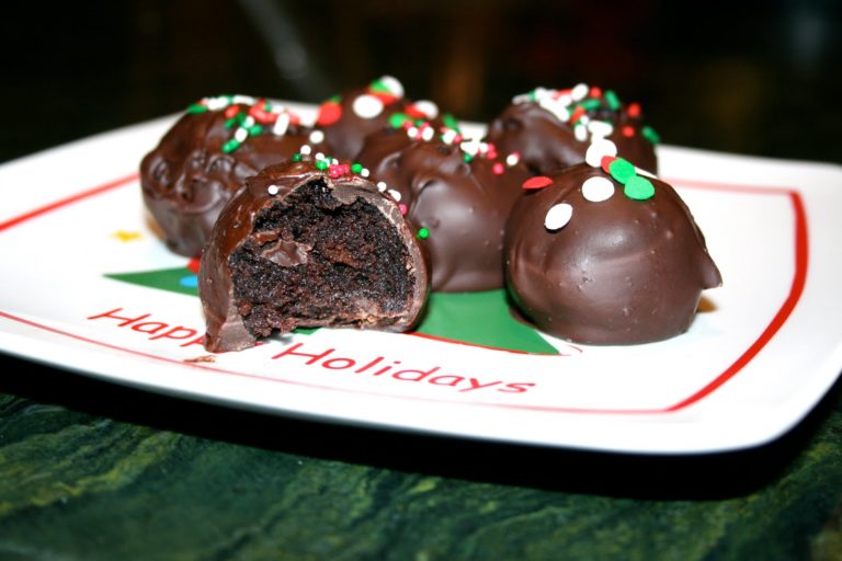 A plate of chocolate cake balls covered in rich chocolate and decorated with festive red, white, and green sprinkles sits atop a “Happy Holidays” plate. One of the chocolate cake balls is bitten into, revealing its moist center.