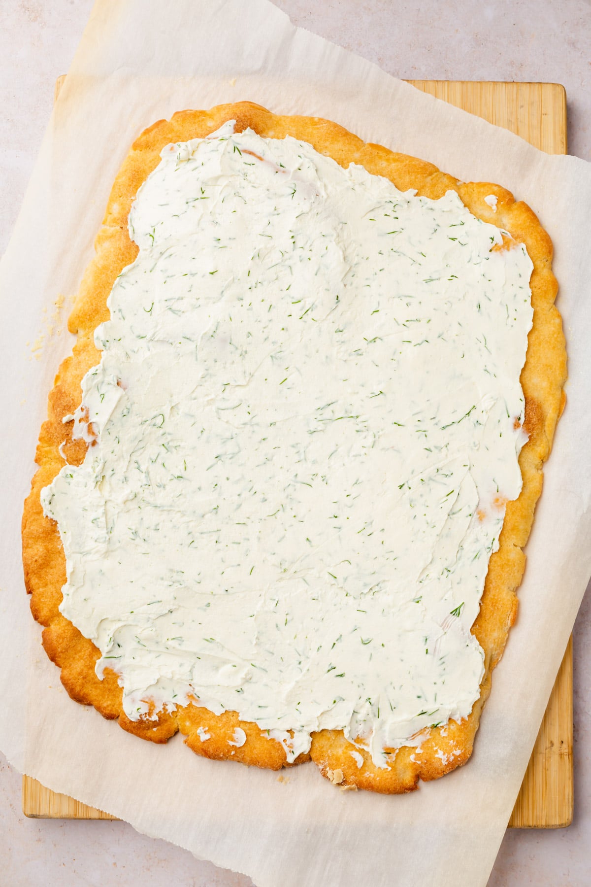 Rectangular gluten-free pizza dough slathered with dill cream cheese on a cutting board lined with parchment paper. 