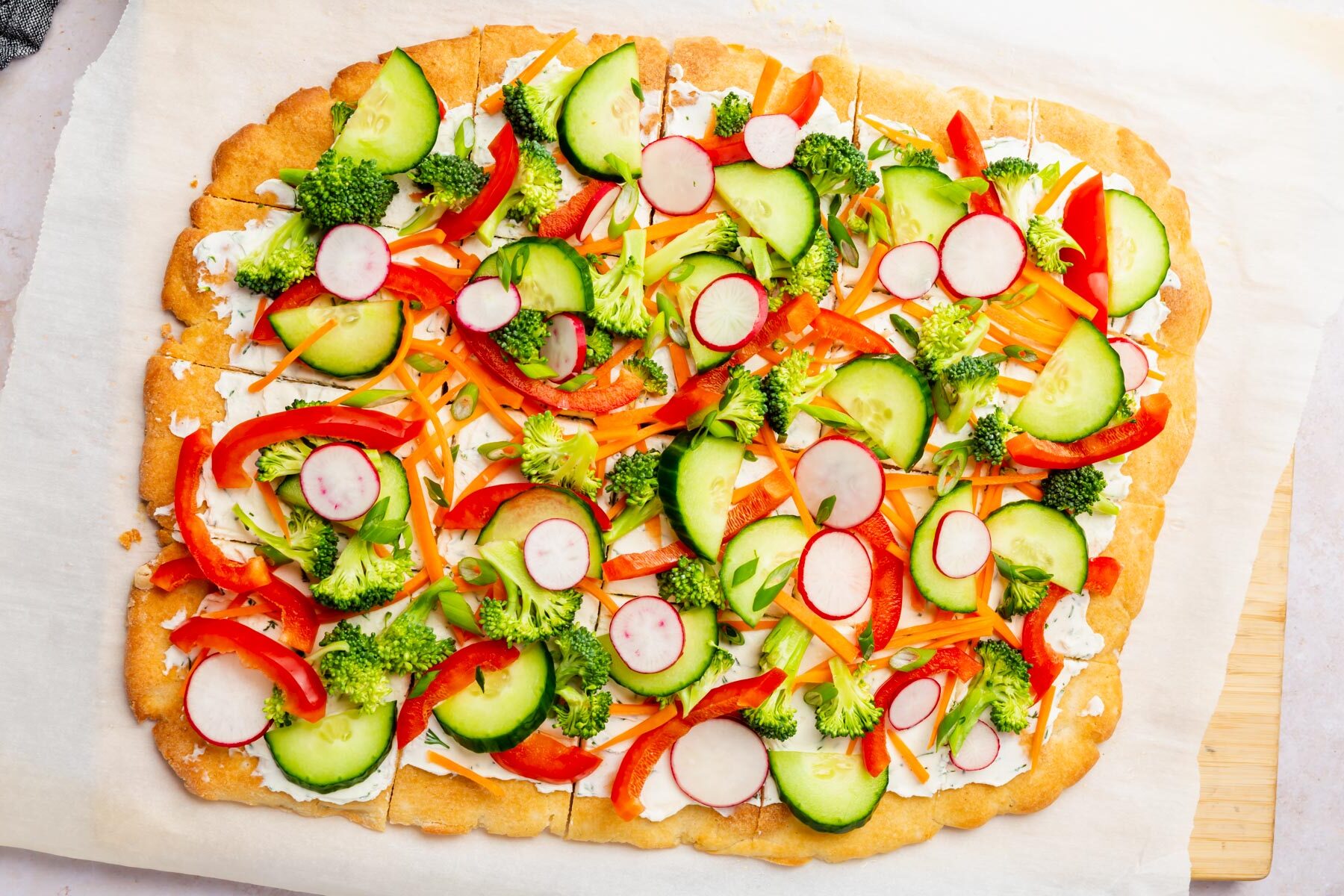 Rectangular gluten-free cold pizza topped with raw cucumber, radish, broccoli, red bell pepper, and shredded carrot.