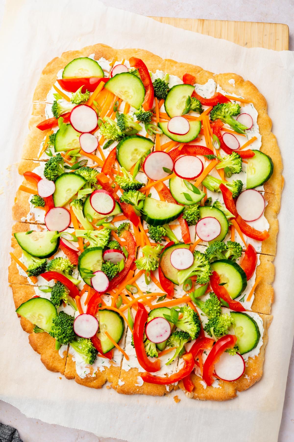 Rectangular gluten-free cold pizza topped with raw cucumber, radish, broccoli, red bell pepper, and shredded carrot.