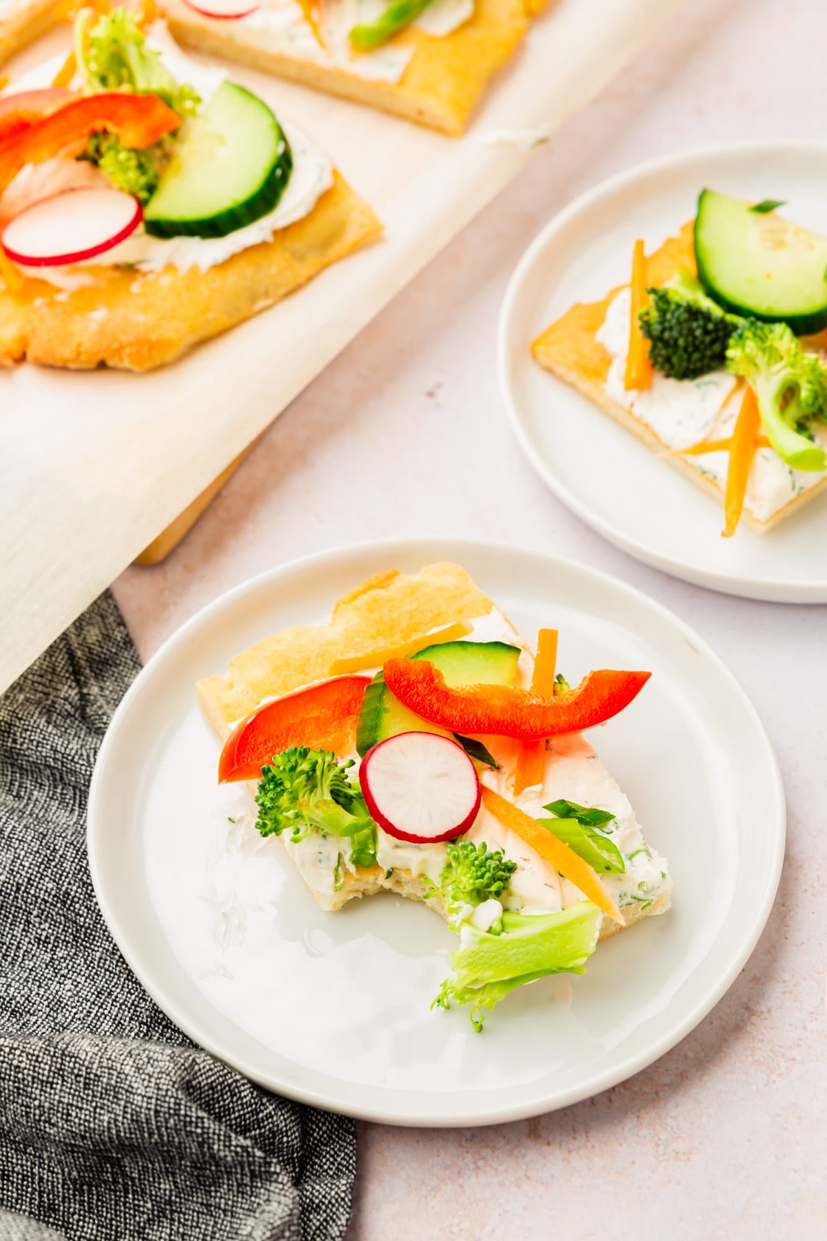 A slice of cold veggie pizza topped with cream cheese, cucumber, radish, bell pepper, broccoli, and shredded carrot sits on a white plate, with extra slices and a gray napkin nearby.