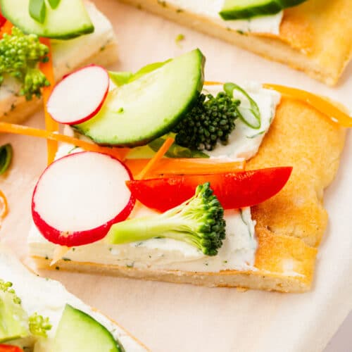 Slices of cold veggie pizza topped with cucumber, radish, bell pepper, broccoli and carrot on a piece of parchment paper.