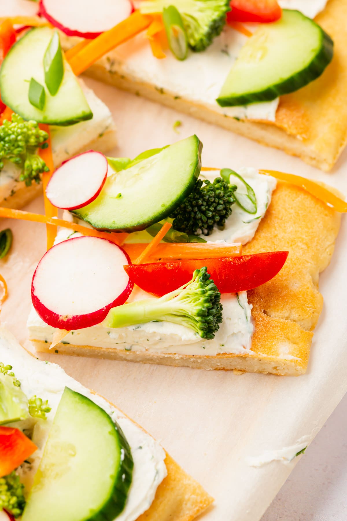 Slices of cold veggie pizza topped with cucumber, radish, bell pepper, broccoli and carrot on a piece of parchment paper.