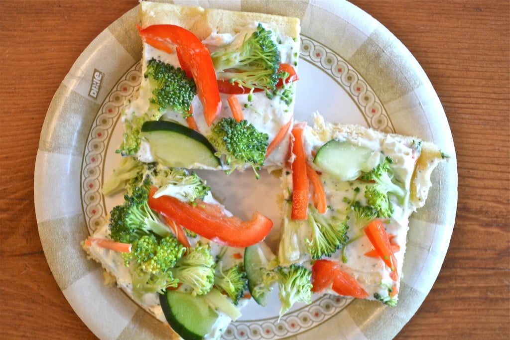 Slices of cold veggie pizza topped with peppers, cucumber and broccoli on a paper plate.