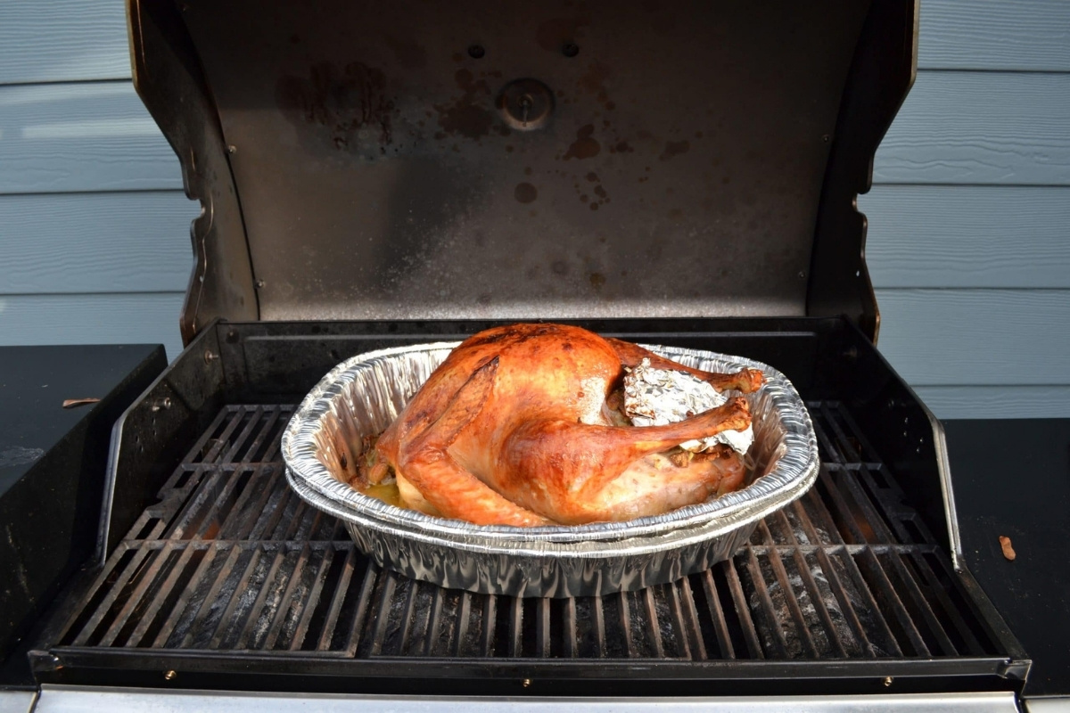 A whole grilled turkey in a foil pan sits on the barbecue grill, golden brown and perfectly stuffed, ready for serving.