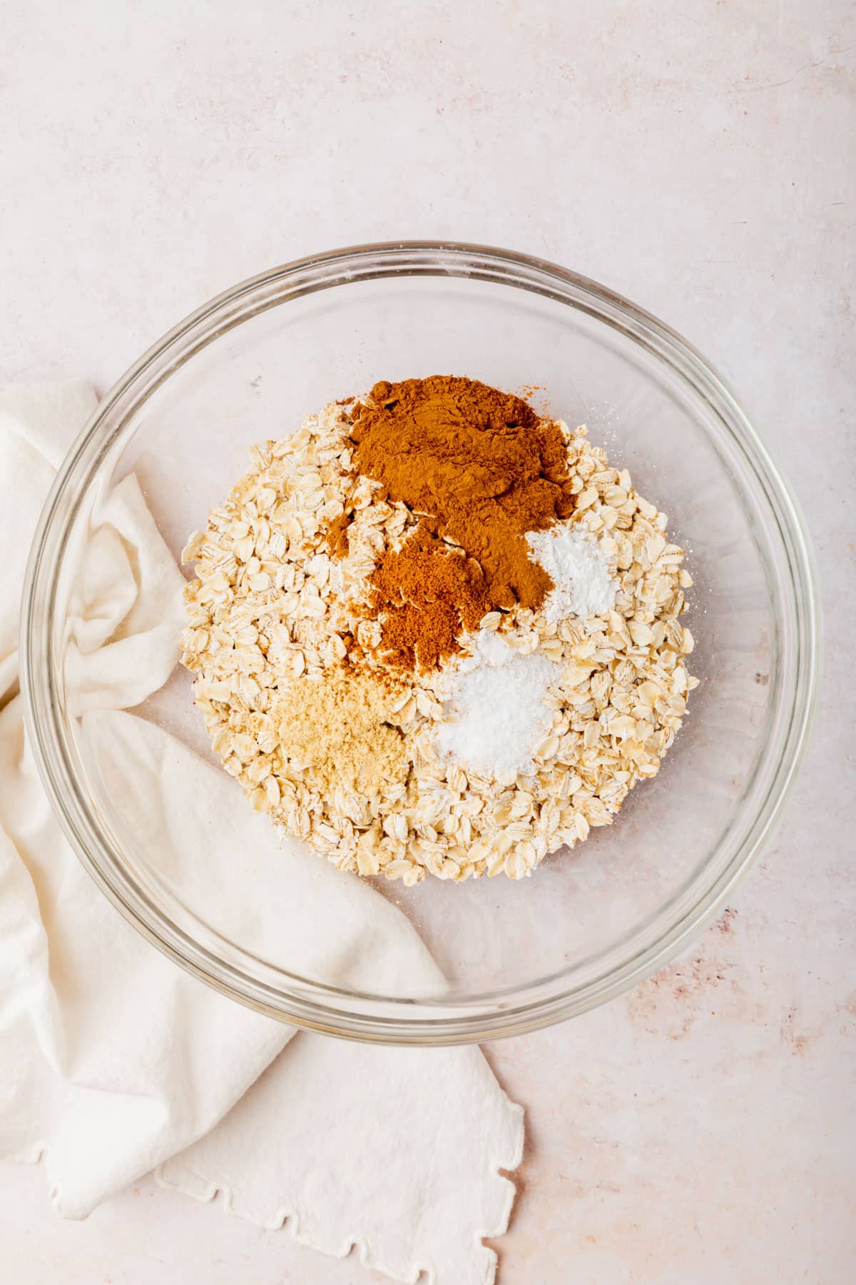 A glass mixing bowl with gluten-free oats topped with cinnamon, nutmeg, ginger, salt, and baking powder before whisking together. 
