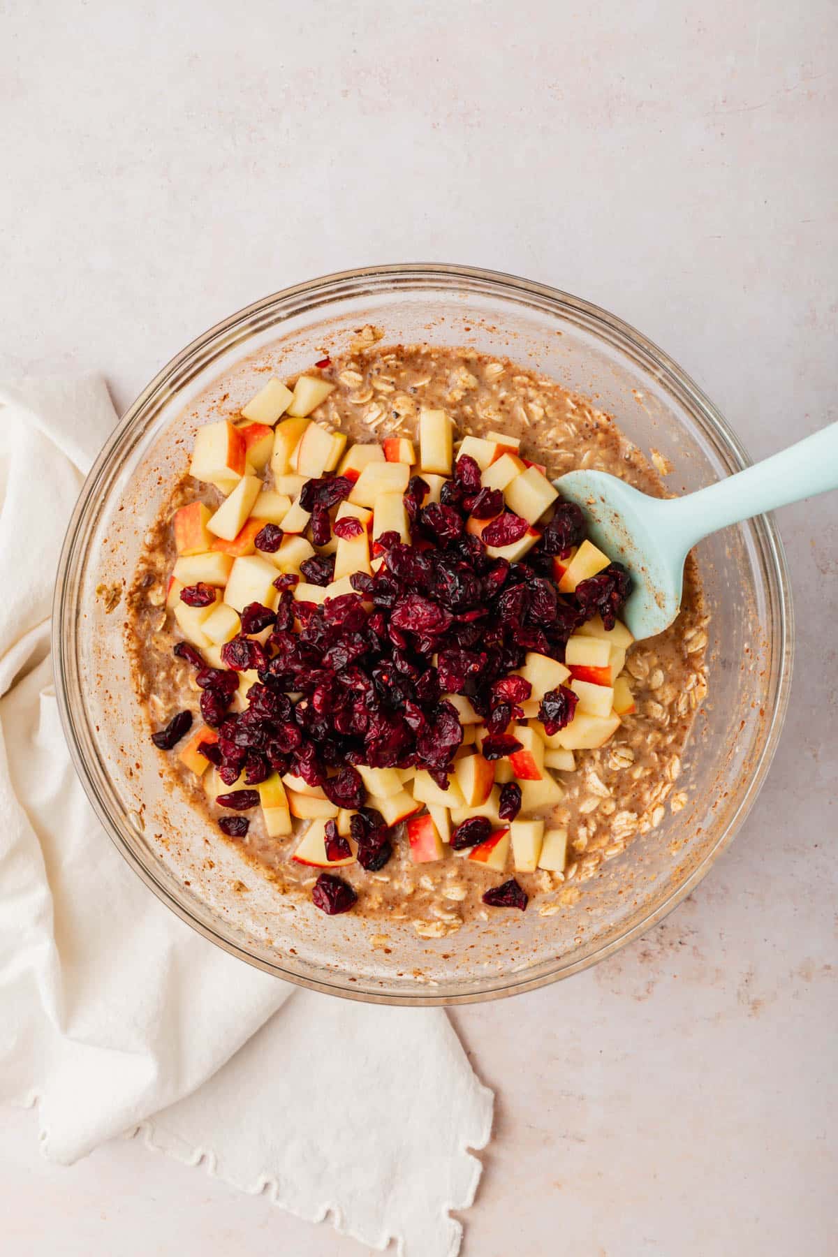 A glass mixing bowl with an oatmeal mixture topped with diced apples and dried cranberries before mixing together.