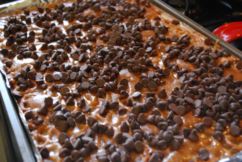 Pretzels lined up on a baking sheet covered with warm caramel sauce and chocolate chips.