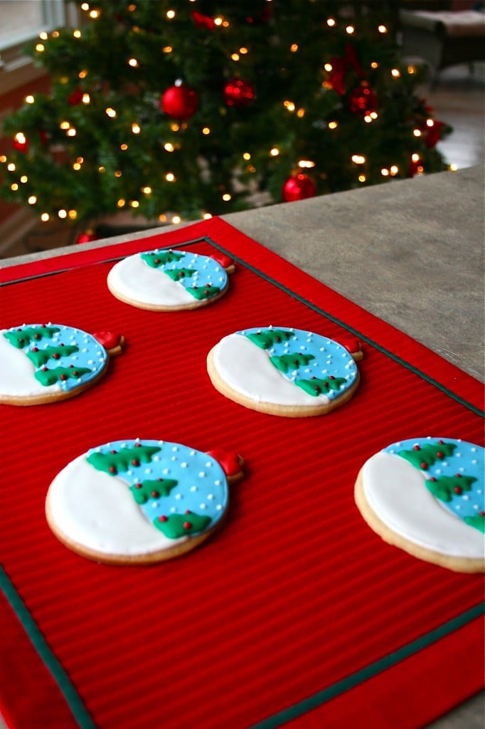 Five snowglobe ornament sugar cookies on a red placemat in front of a Christmas tree.