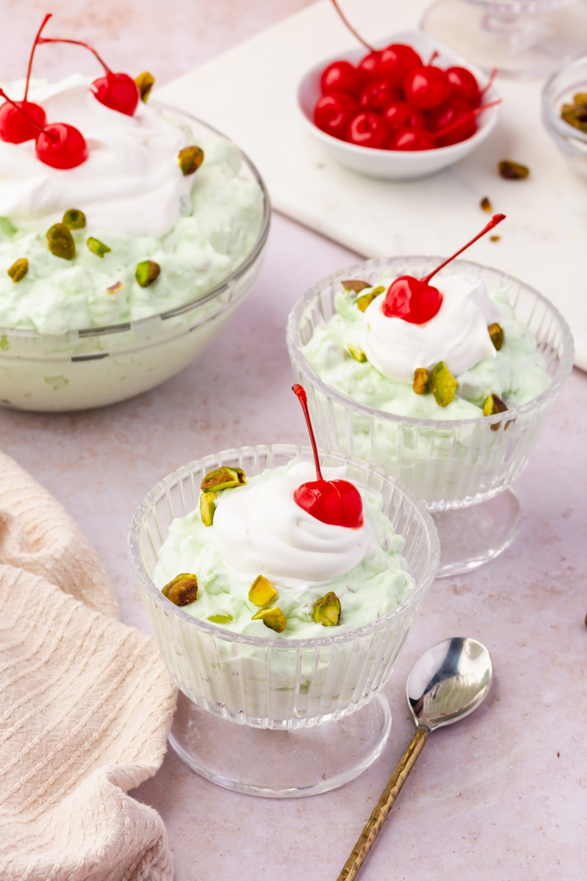 Two glass bowls and a larger dish filled with Watergate Salad, a light green dessert topped with whipped cream, pistachios, and a maraschino cherry. Spoons and a small plate of cherries are nearby on a light surface.