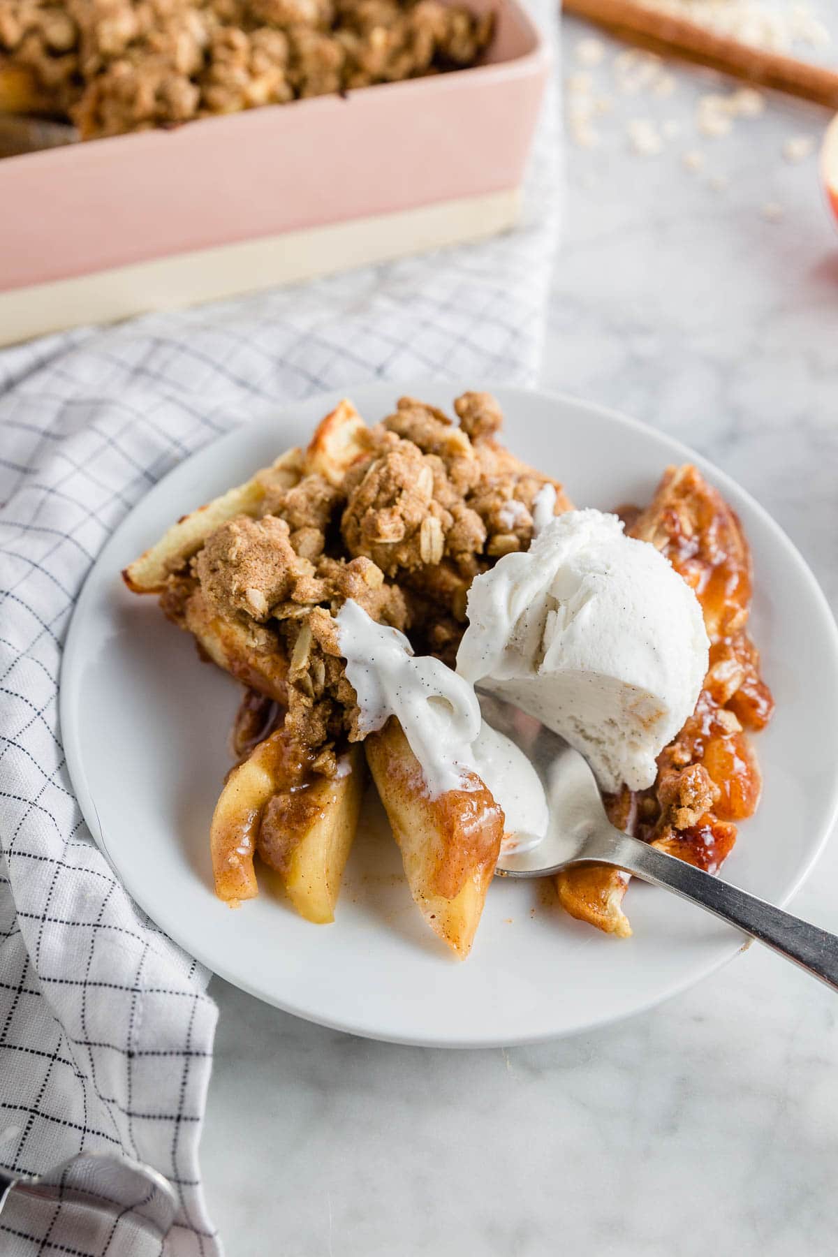 A white plate with gluten-free apple crisp with a scoop of melty vanilla ice cream with a spoon digging in. 