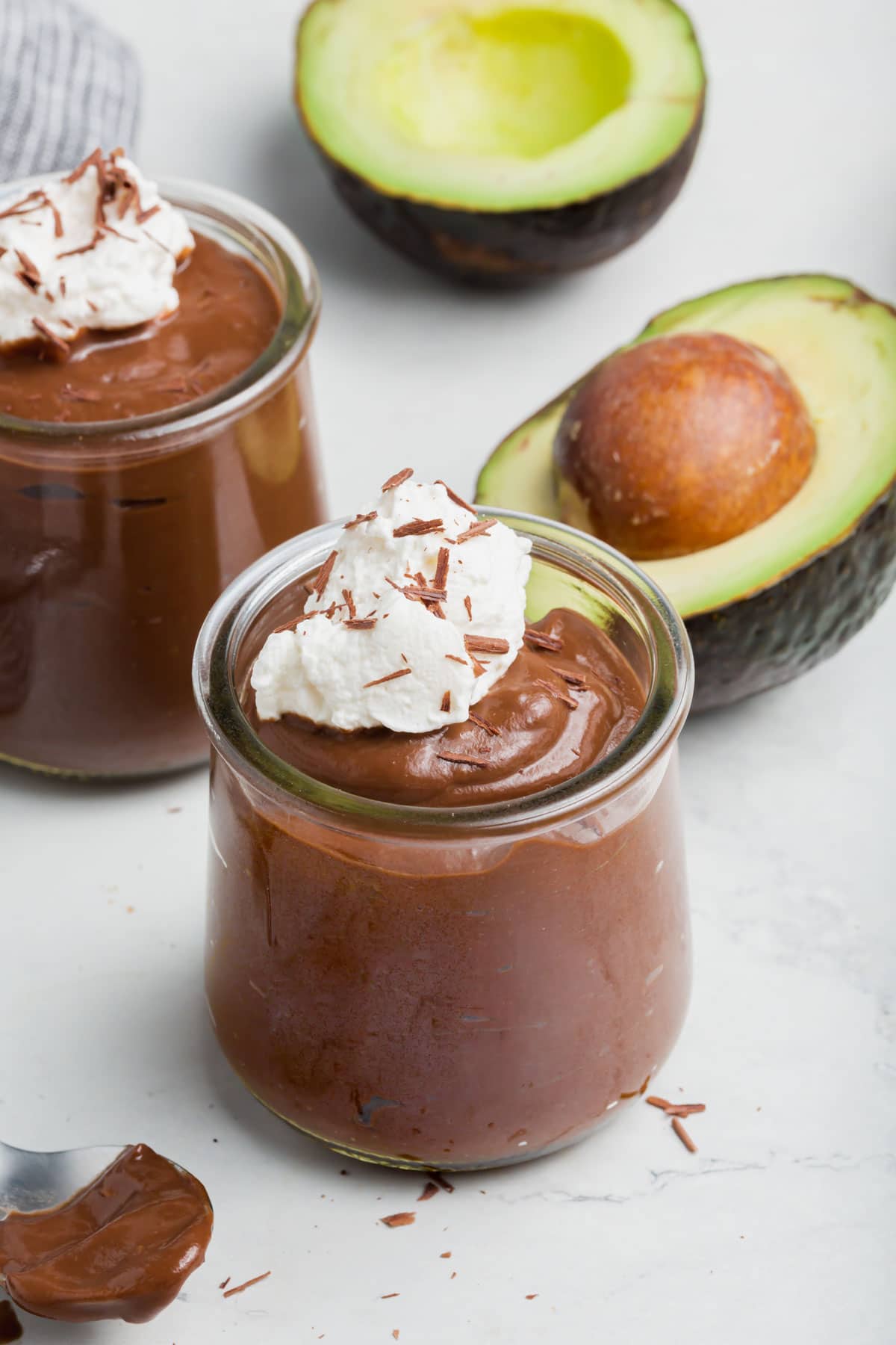 Two jars of chocolate avocado pudding with coconut whipped cream on top with avocados in the background.