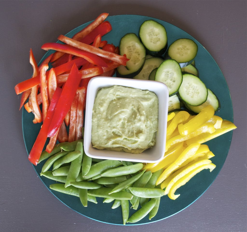 A bowl of avocado bean dip in the center of a platter of cucumber, yellow bell peppers, snap peas, and red bell peppers.