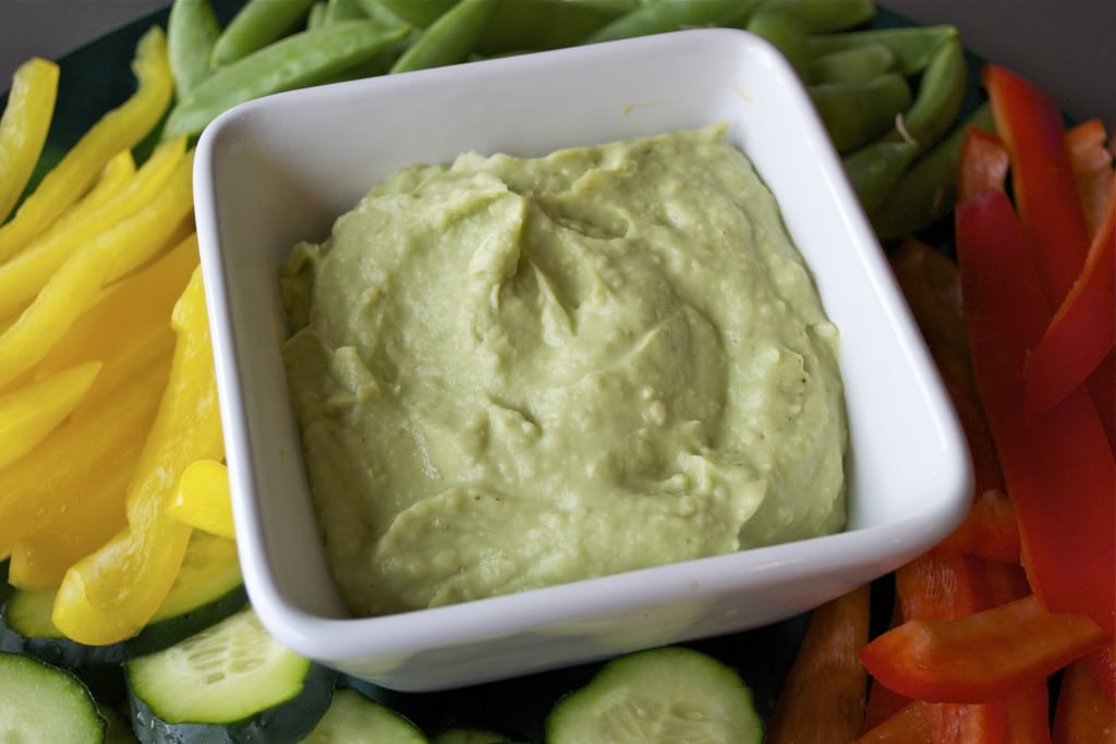 A bowl of avocado bean dip surrounded by bell peppers, cucumbers and snap peas.