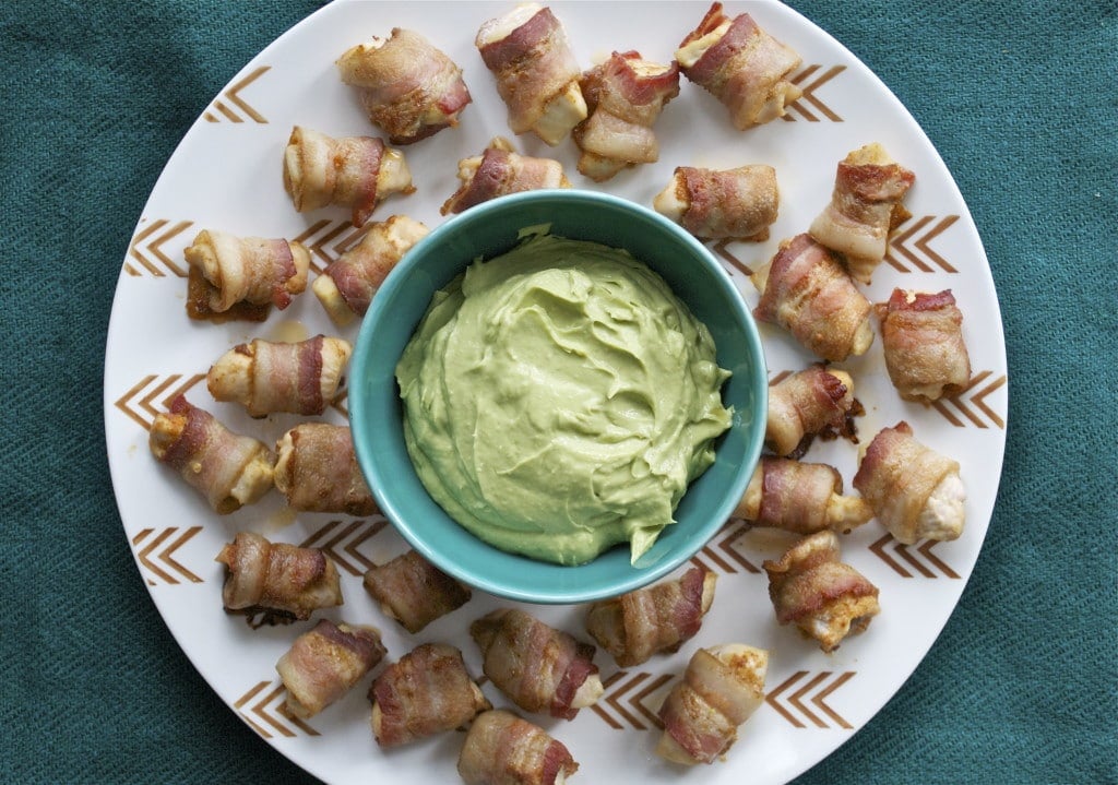 A platter of bacon wrapped chicken bites surrounding a bowl of avocado dipping sauce.