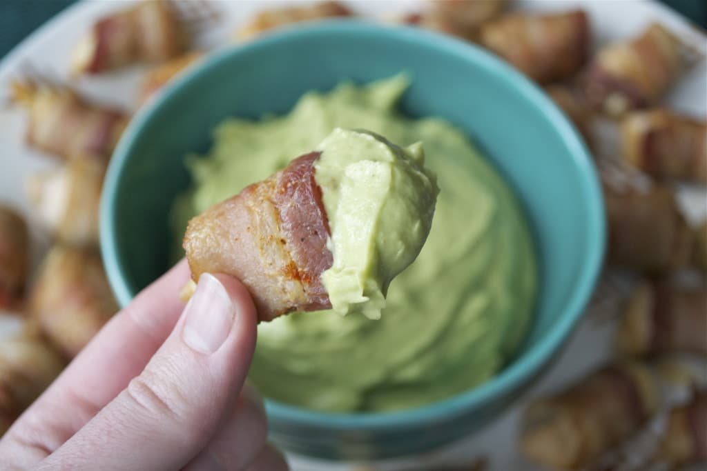 A hand holding a bacon wrapped chicken bite dipped in avocado dipping sauce.