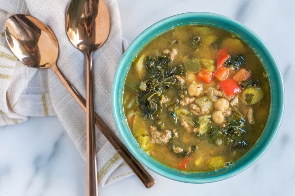 A bowl of turkey soup with kale, peppers, chickpeas and brussels sprouts next to two spoons. 