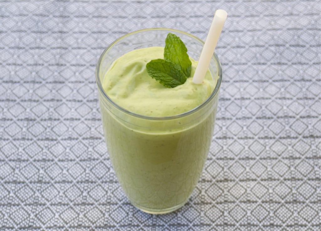 A green shake in a glass topped with fresh mint leaves and a straw.