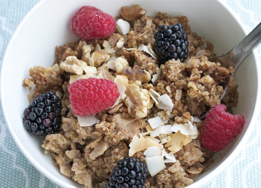 A bowl of cinnamon baked quinoa topped with walnuts, toasted coconut, raspberries and blackberries with a spoon in it.