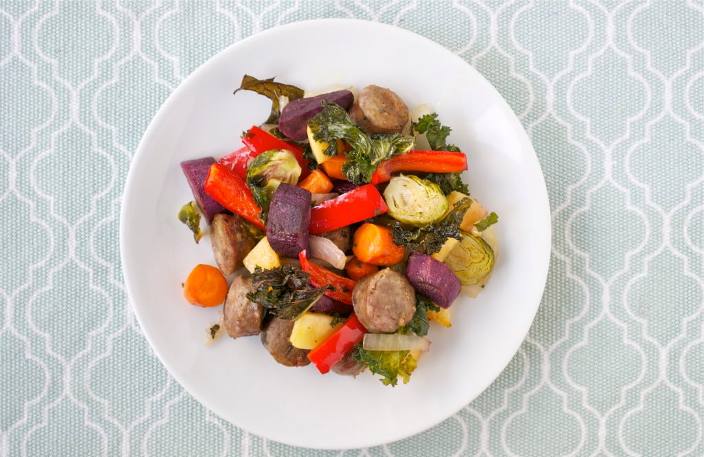 Roasted sausage, purple sweet potato, red bell peppers, brussels sprouts, carrots and kale on a white plate. 