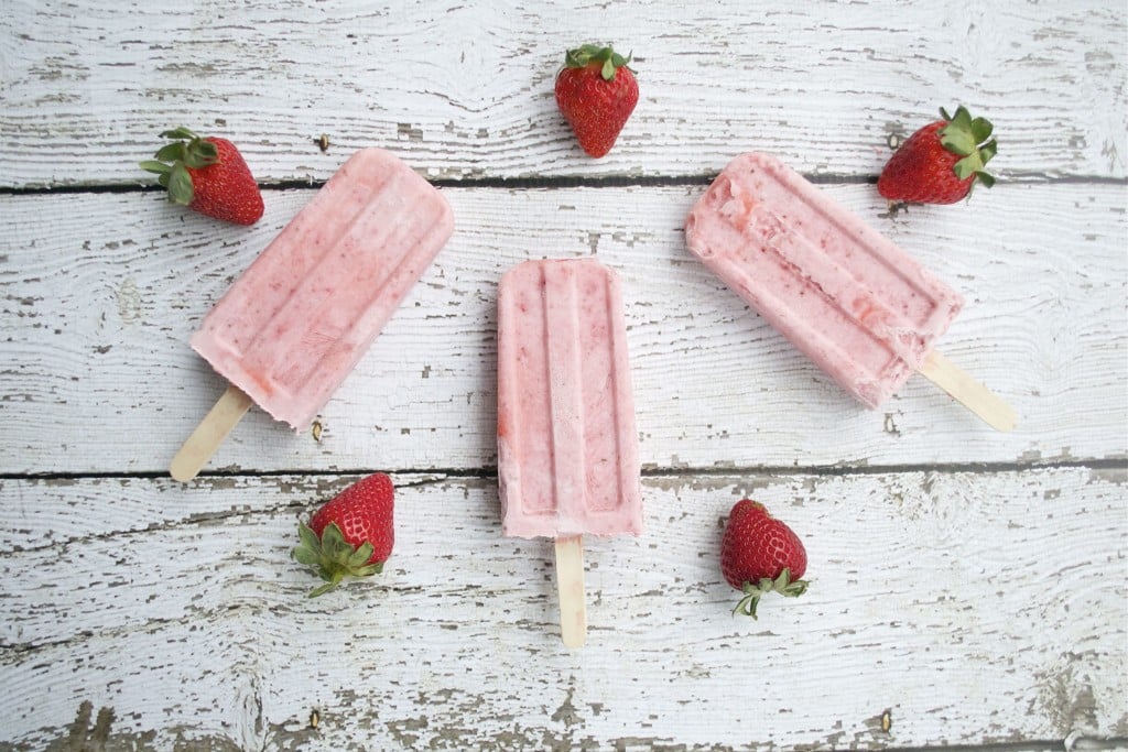 Three strawberry and cream popsicles on a white wood table with fresh strawberries.