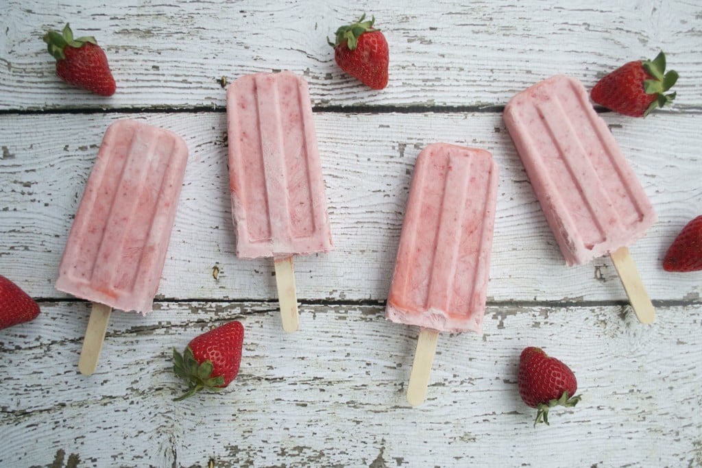 Four strawberry and cream popsicles on a white table with fresh strawberries.