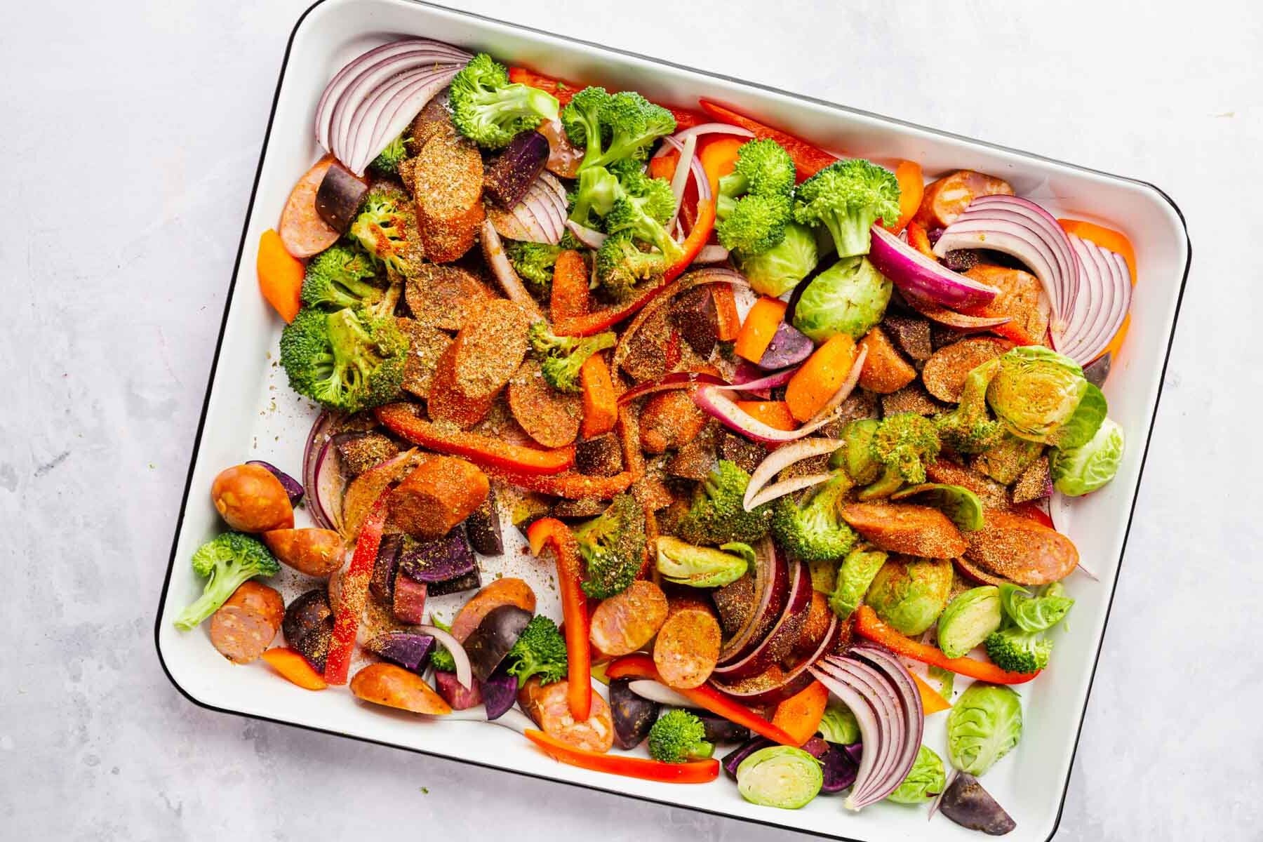 Sausage, vegetables and spice mixture in a white sheet pan before baking.