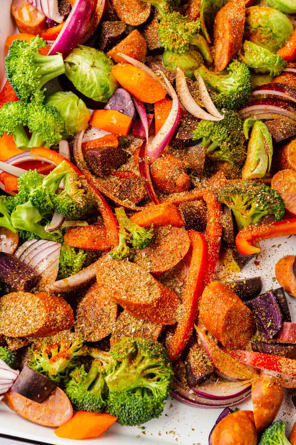 A close up of sausage, vegetables and spices on a white sheet pan.