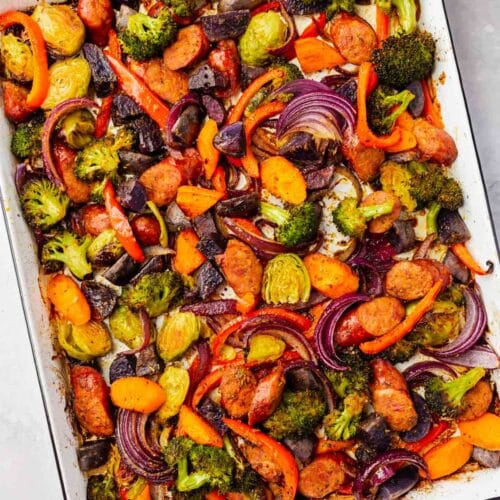 An overhead view of roasted sausage and vegetables on a white sheet pan after baking.