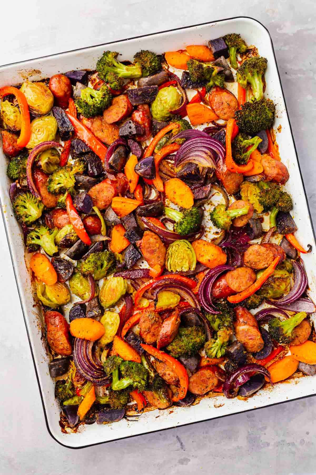 An overhead view of roasted sausage and vegetables on a white sheet pan after baking.