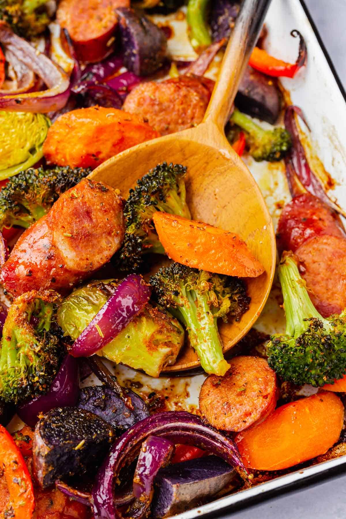 A wood spoon scooping up roasted sausage and vegetables from a sheet pan.