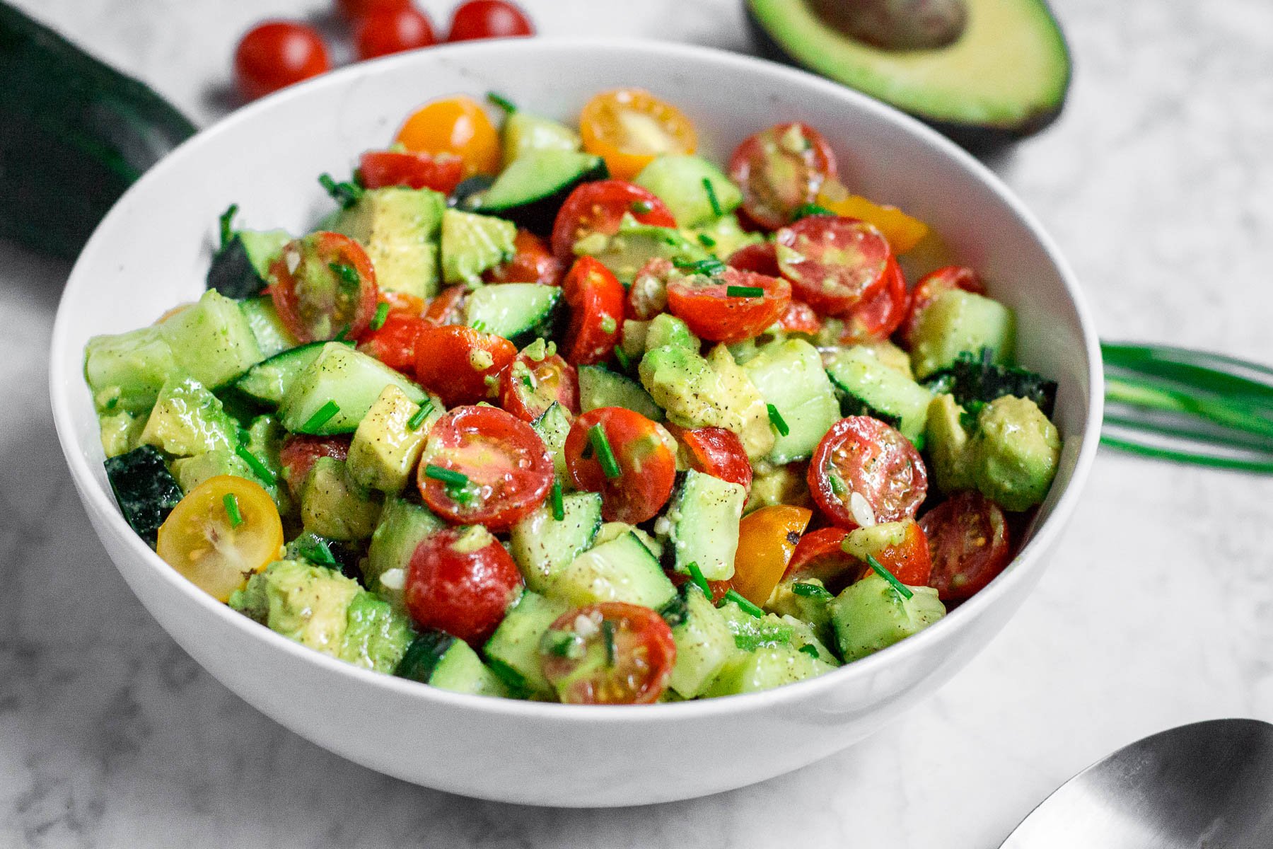 A white bowl filled with diced cucumbers, tomatoes, and avocado in a lemon dressing with chopped chives on top. 