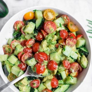 An aerial view of a white bowl filled with cucumber tomato avocado salad with a spoon ready for serving.