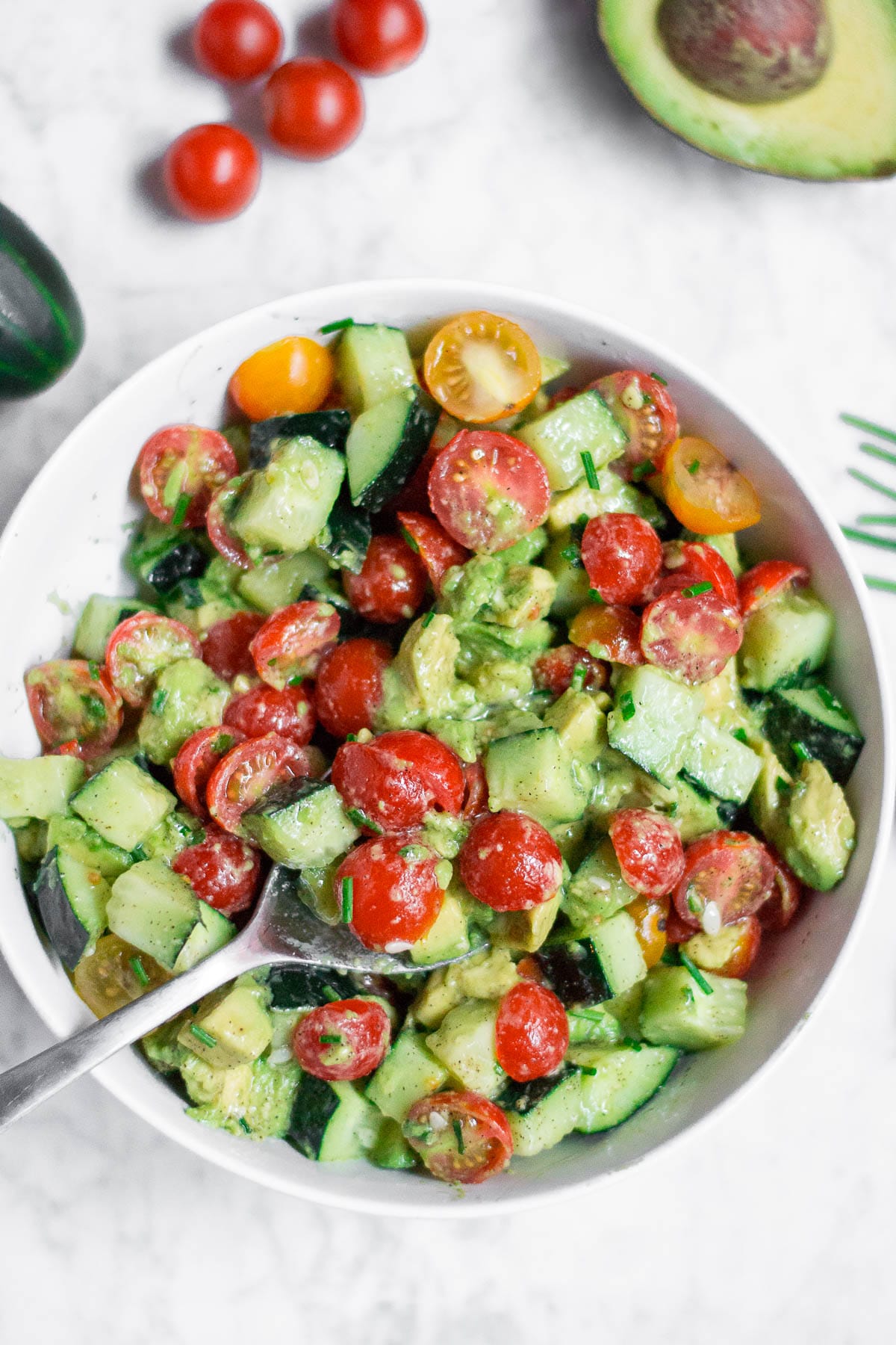 An aerial view of a white bowl filled with cucumber tomato avocado salad with a spoon ready for serving. 