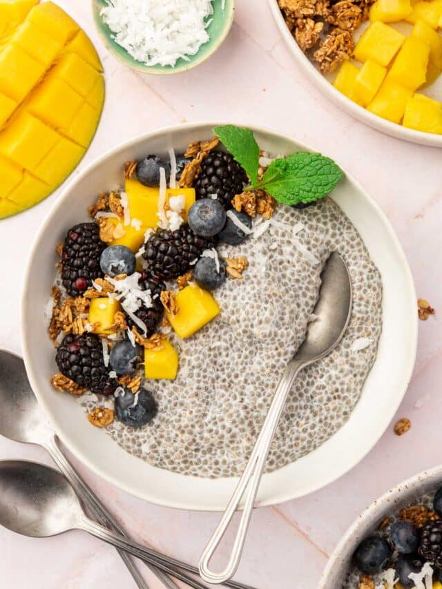 A bowl of chia pudding topped with mango, blueberries, blackberries, coconut and granola with more toppings surrounding the bowl.