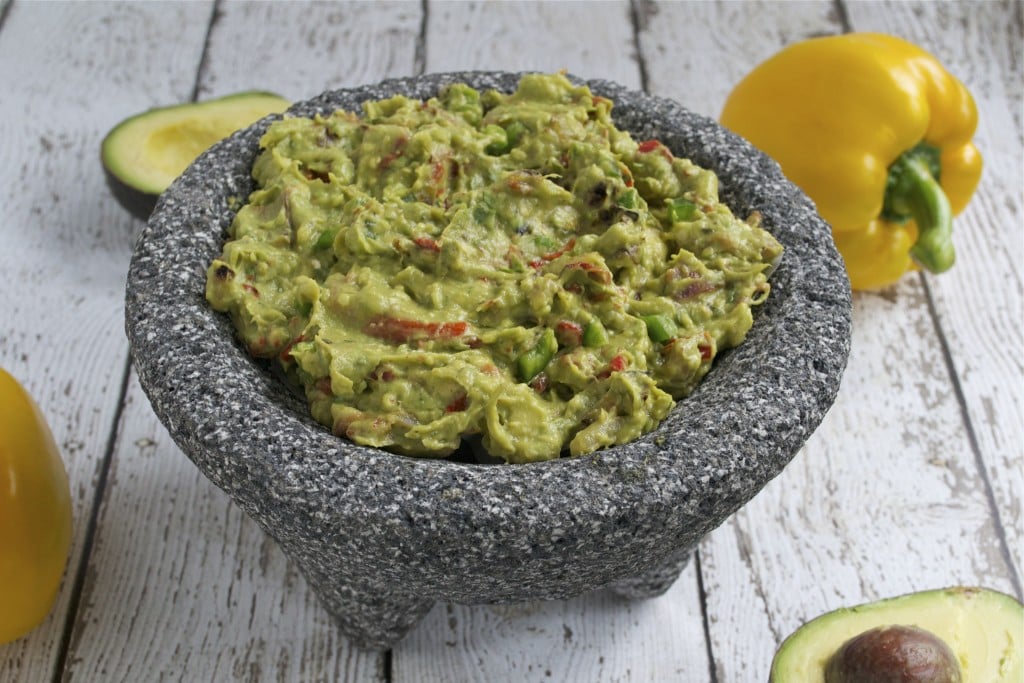 A molcajete filled with guacamole with bell peppers and avocados in the background.