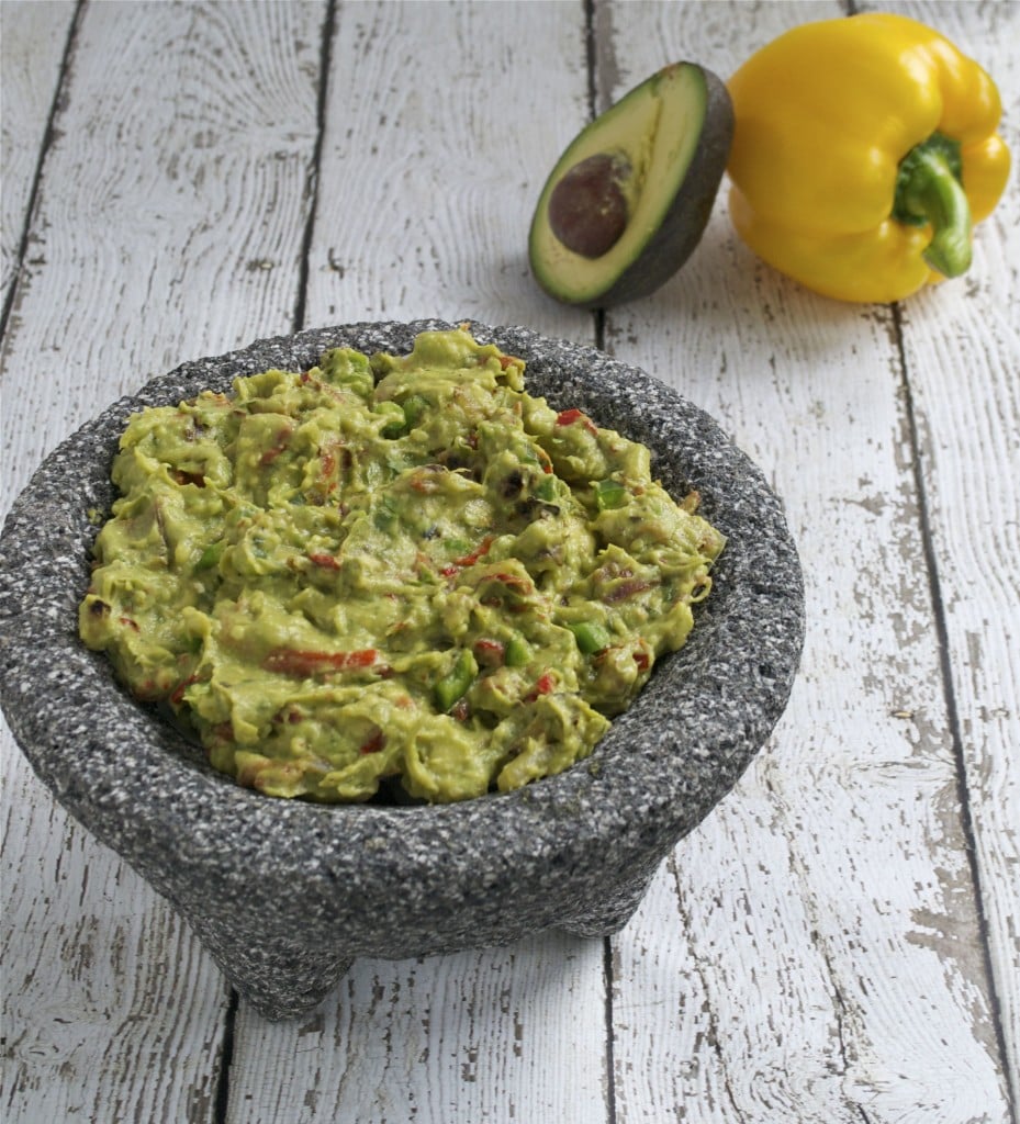 A molcajete filled with bacon and roasted bell pepper guacamole with an avocado and bell pepper in the background.