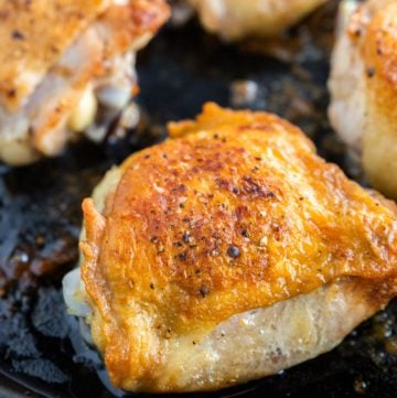 A photo of a cast iron skillet with crispy roasted chicken thighs topped with salt and pepper in it.