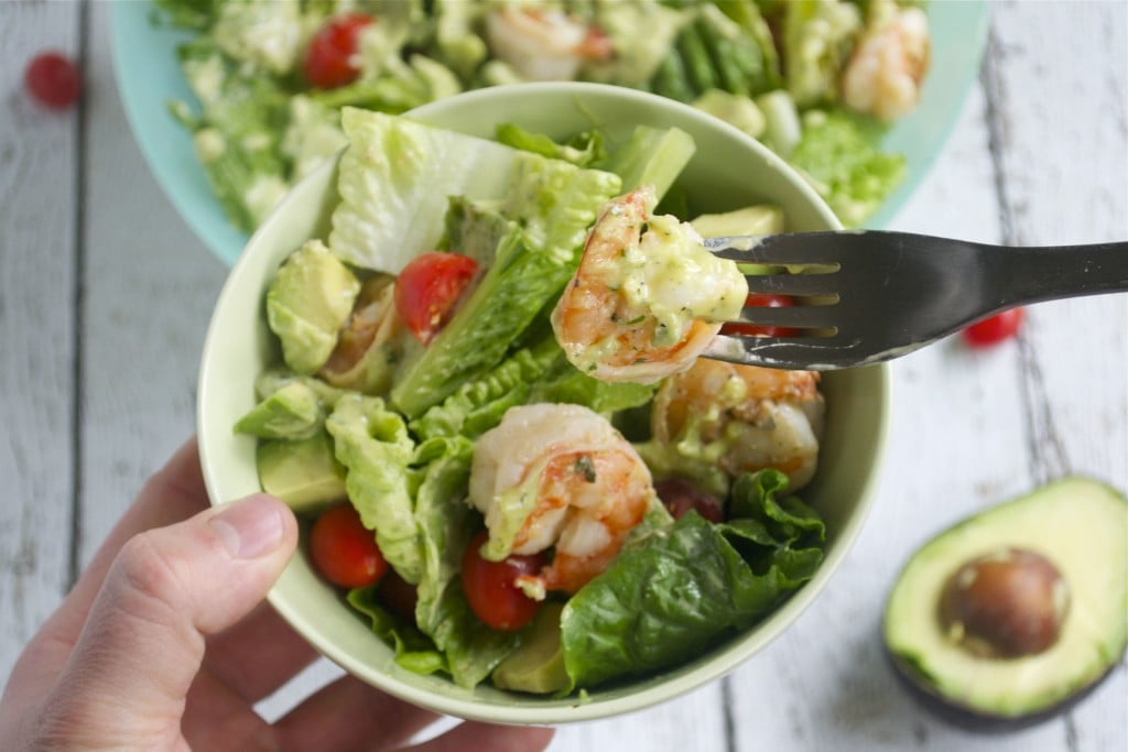 A hand holding a bowl of shrimp salad with cherry tomatoes and avocado with a fork digging in.