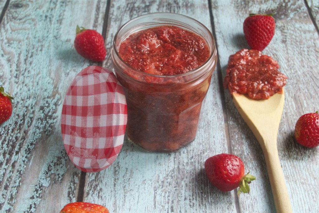 A jar with strawberry chia jam with a checkered lid and a wooden spoon with jam on it. 