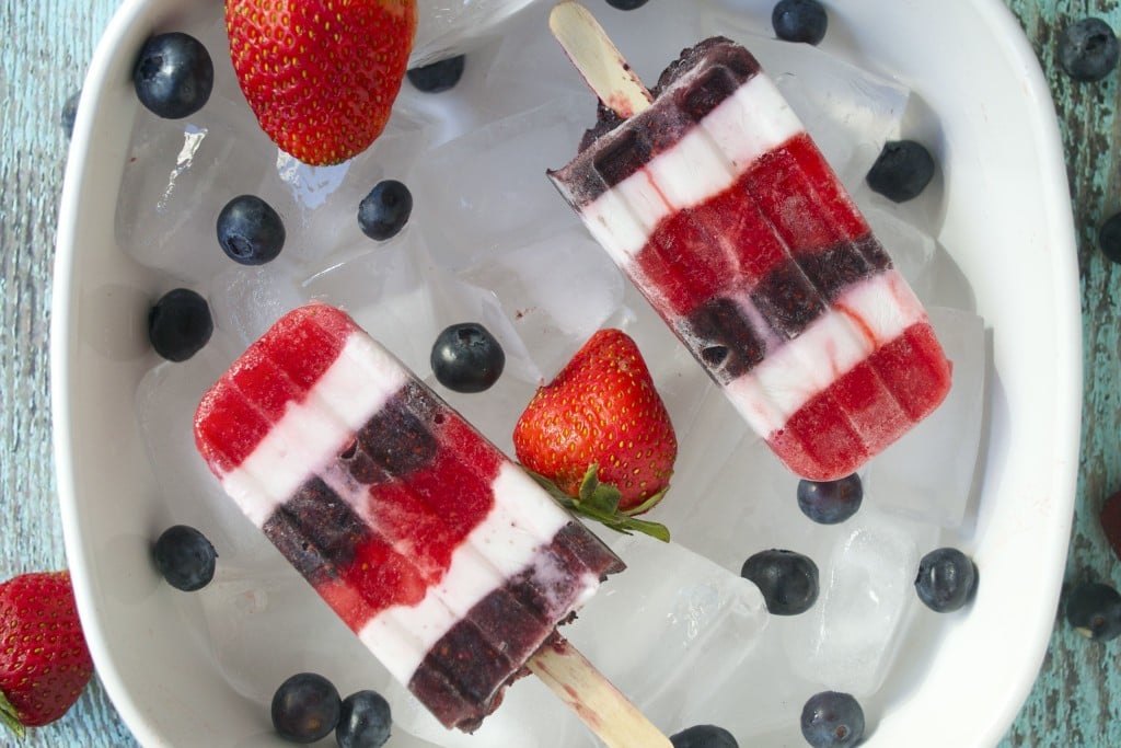 A bowl of ice topped with two layered red, white and blue popsicles surrounded by fresh blueberries and strawberries.
