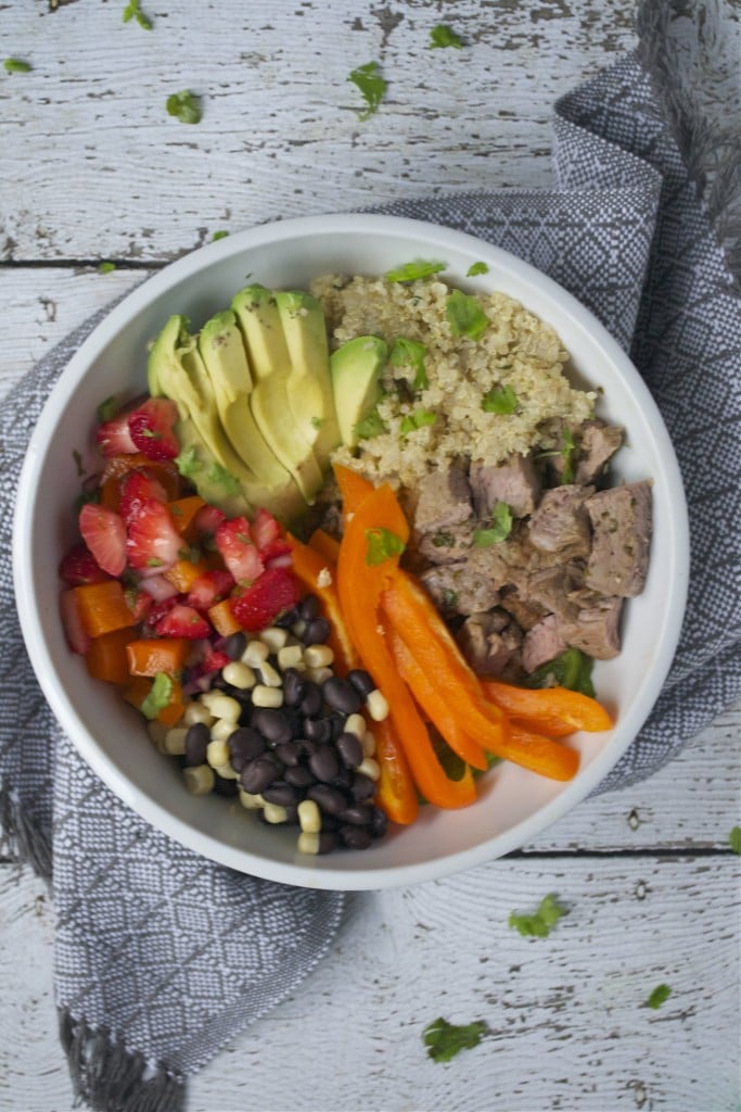 An aerial view of a white bowl with steak, quinoa, avocado, strawberries, black beans, corn, bell peppers on a white wood table with a grey linen napkin.