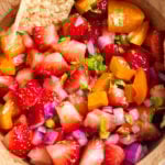 Close-up of strawberry salsa in a wooden bowl with a tortilla chip, featuring chopped strawberries, orange bell pepper, red onion, jalapeño, and cilantro.